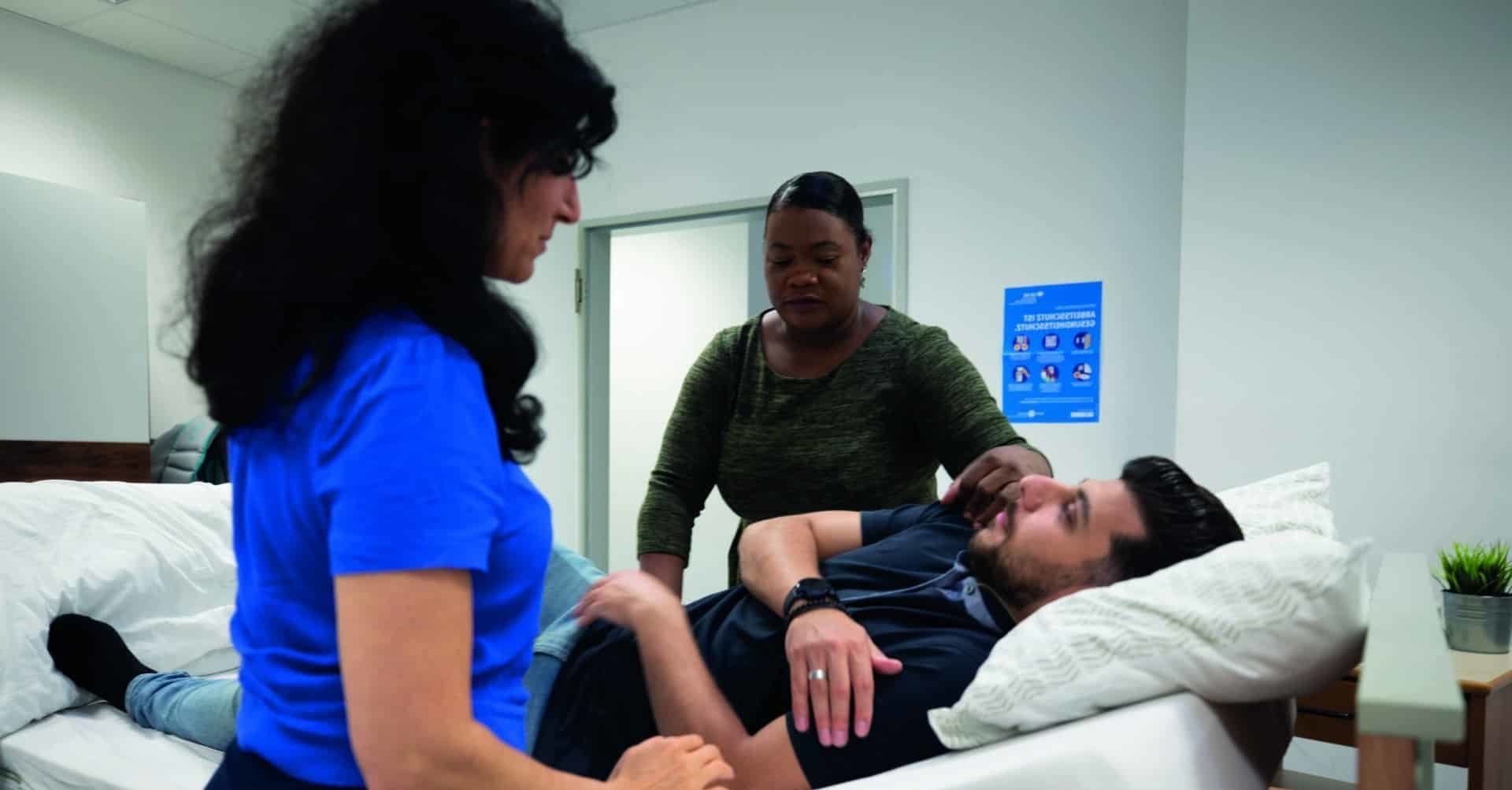 Eine Fachkraft demonstriert einen Behandlungsvorgang beispielhaft an einem Lerndenden auf einem Krankenbett liegend.